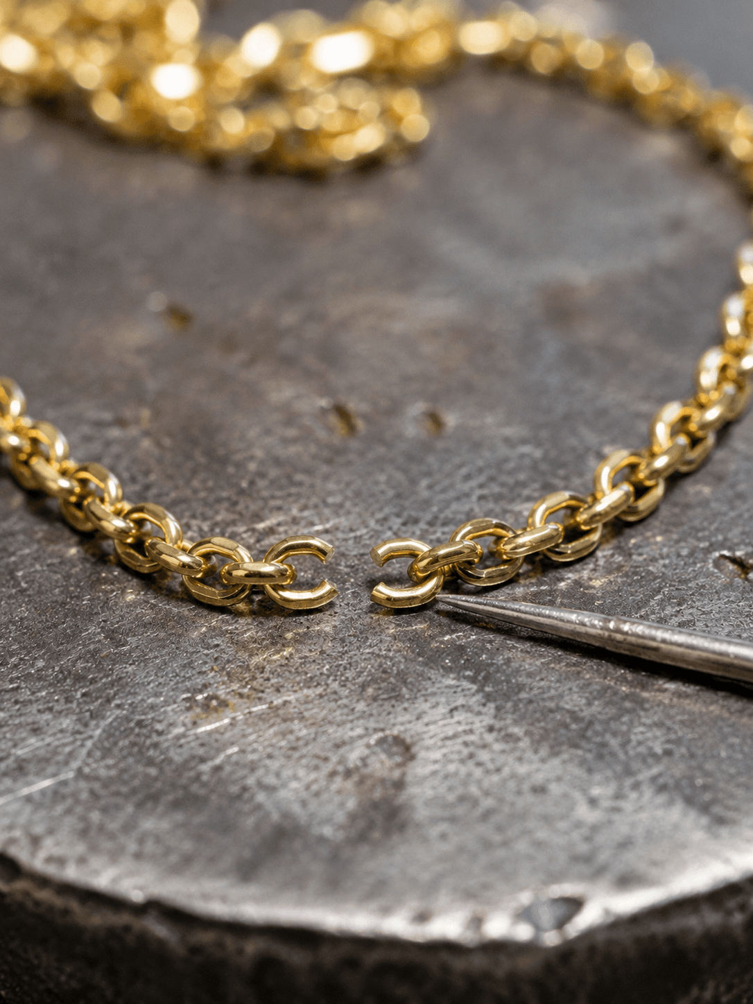 Gold rolo chain with an open link beside a jeweler's steel pick on a bench block