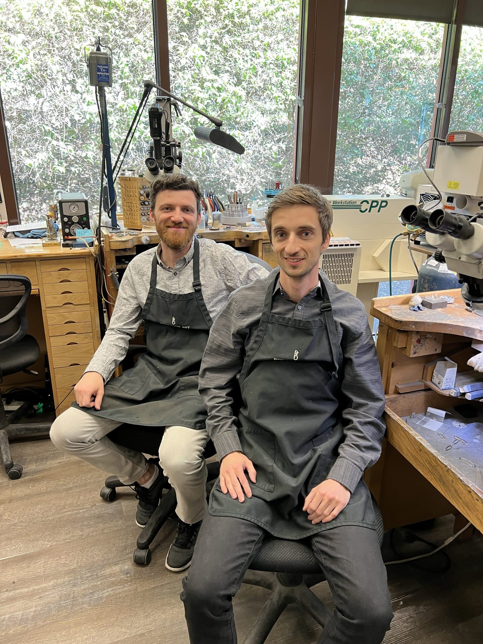 Two jewelers in dark aprons seated at jewelry workbench with microscope and tools, workshop setting.