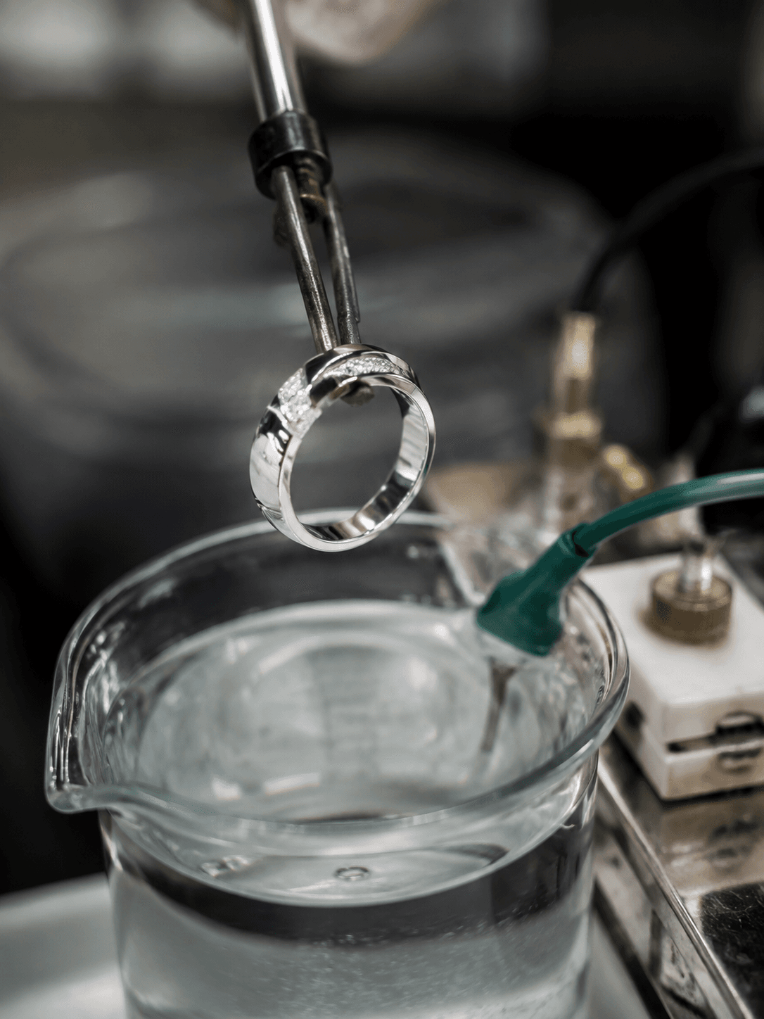 White gold diamond ring held by metal tongs above a glass beaker of plating solution