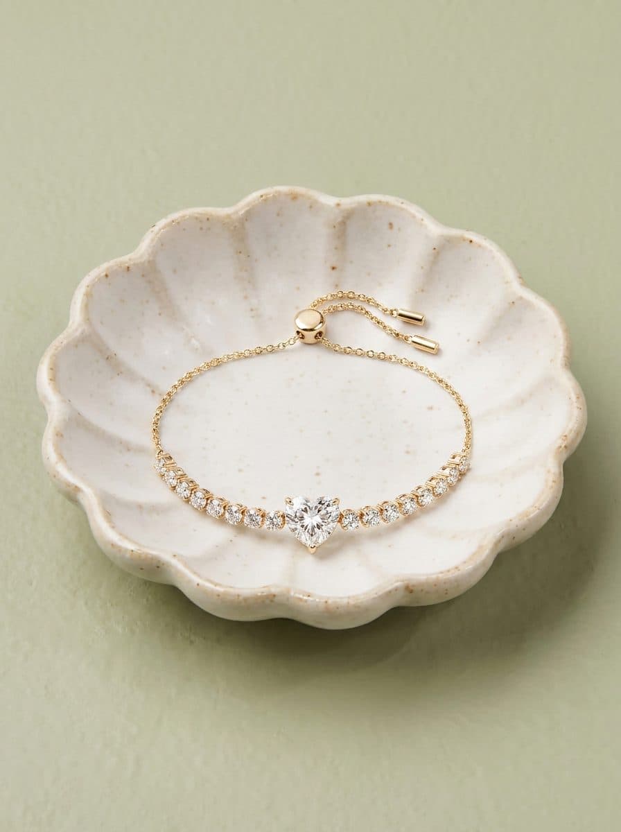 Gold-tone adjustable tennis bracelet, heart-cut CZ center, round CZ accents, slider clasp, scalloped ceramic dish, sage