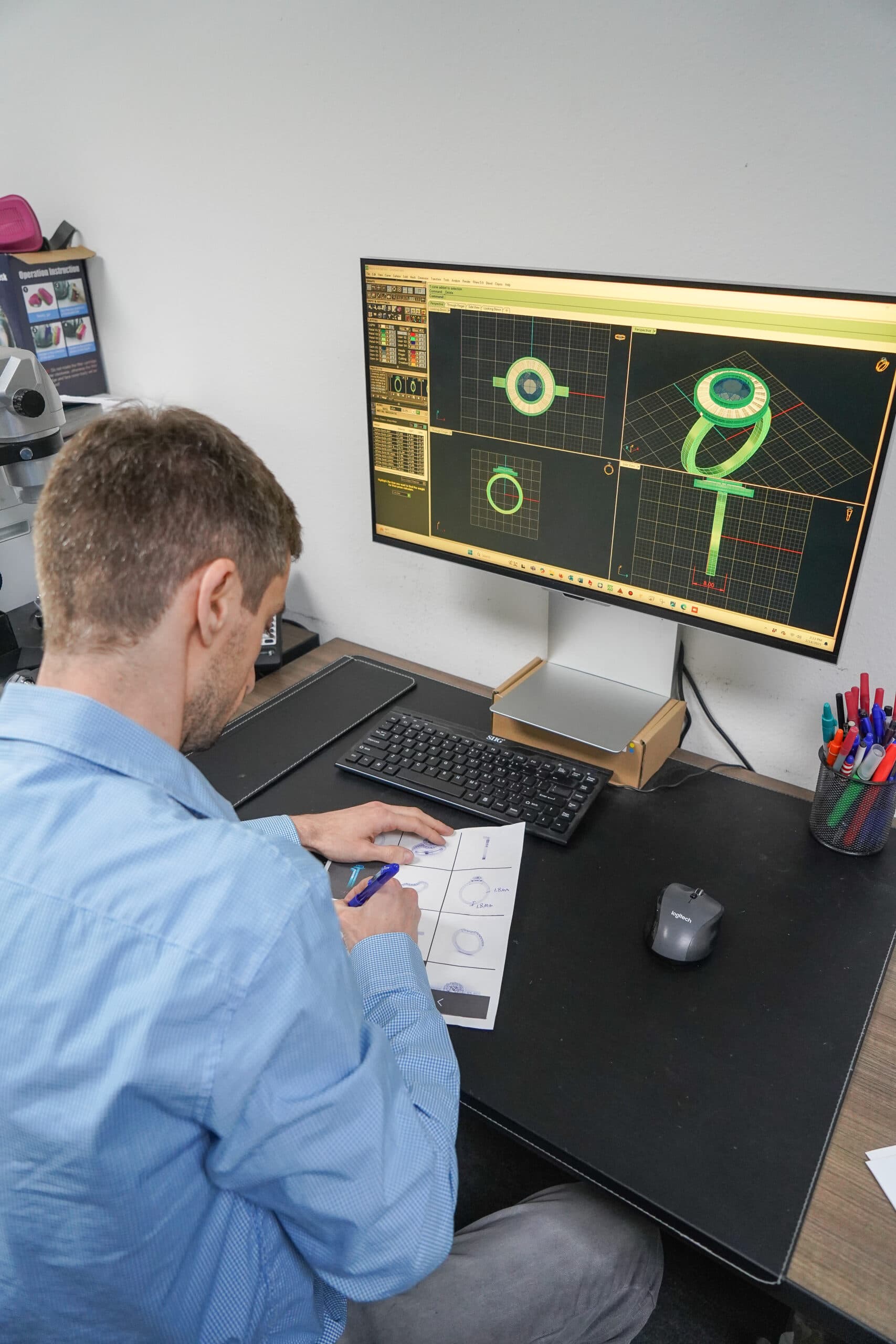 Jewelry designer reviewing 3D CAD ring model on monitor with printed sketches at studio workstation.