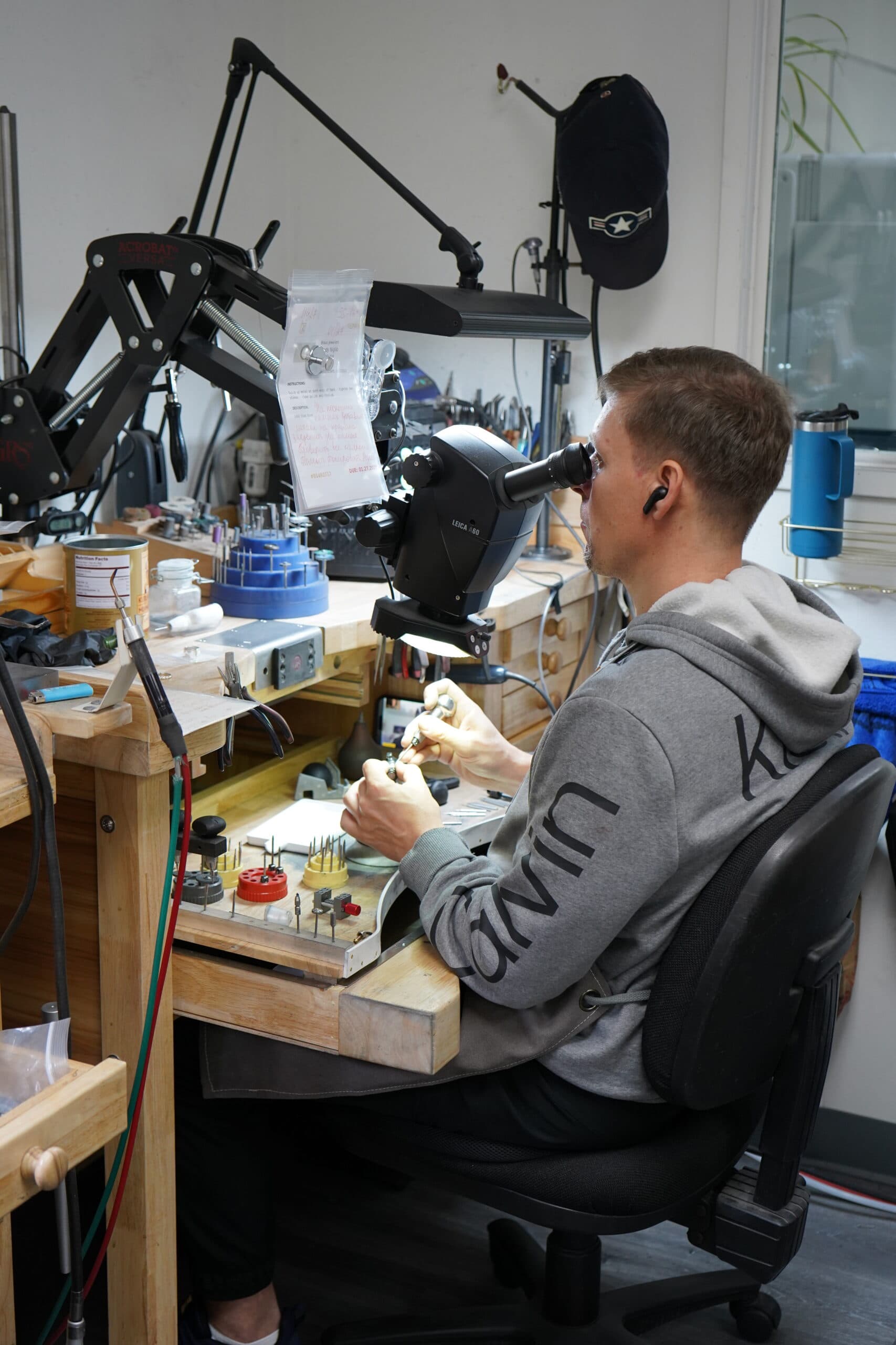 Jeweler working at bench using Leica microscope, jewelry workshop setting with tools and burr bits.