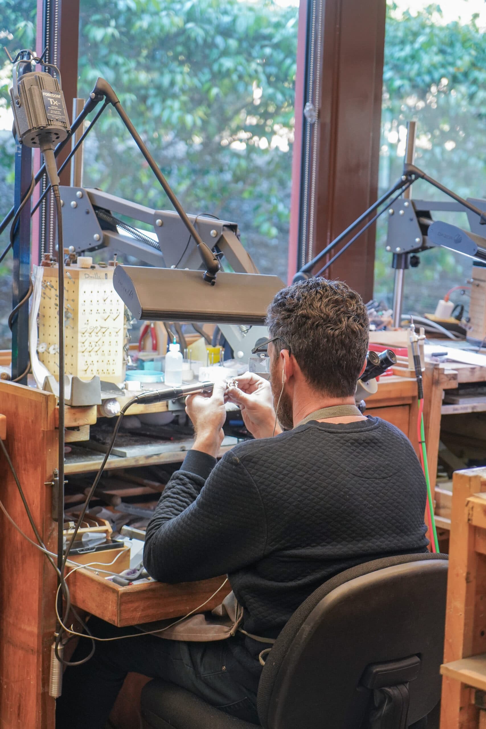 Male jeweler working at bench with tools, flex shaft drill, task lighting; workshop setting, window background.
