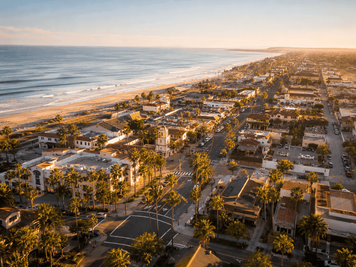 Aerial view of coastal California town with palm trees, Spanish-style buildings, sandy beach, and ocean at golden hour.