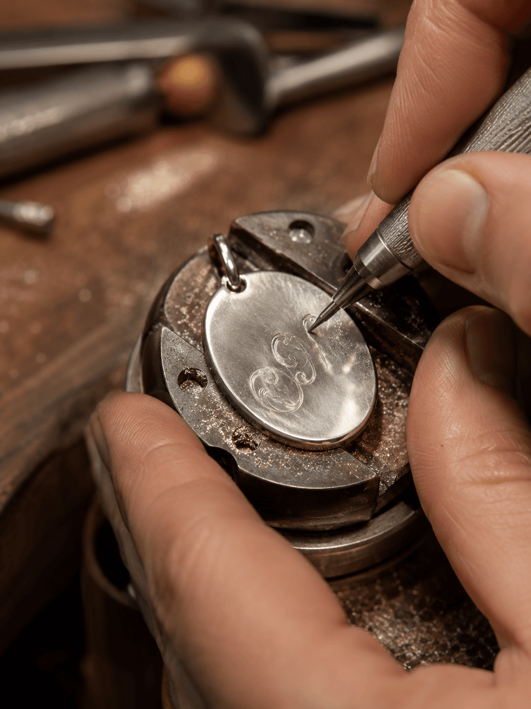 Jeweler hand-engraving a monogram onto a polished oval silver pendant held in a bench vise